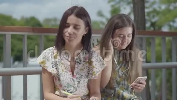 两个漂亮的女学生坐在户外一个女孩带着耳机听音乐第二个女孩视频的预览图
