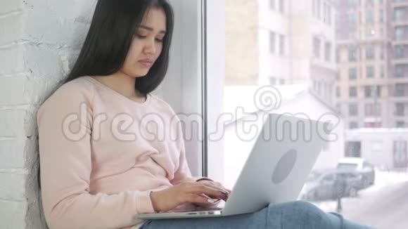 穿着笔记本电脑工作的美国年轻女性从窗户看视频的预览图