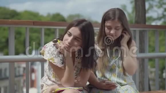 两个漂亮的女学生坐在户外一个女孩带着耳机听音乐第二个女孩视频的预览图