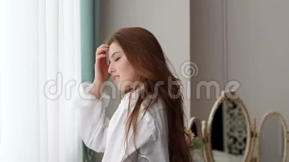 穿着白色衬衫的性感红发女孩站在卧室的大窗户附近视频的预览图