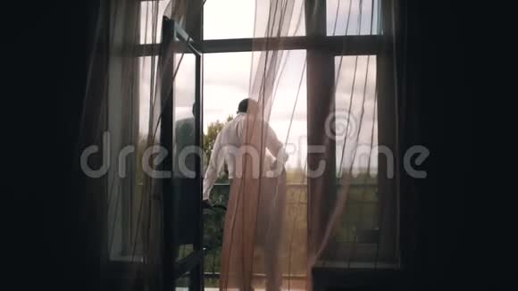 时髦的年轻人穿上夹克站在阳台上从房间里透过门帘看到美丽的景色视频的预览图