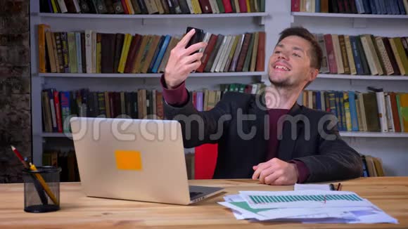 有魅力的男学生在图书馆里坐在笔记本电脑前打电话自拍视频的预览图