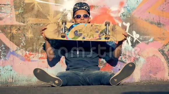 一个微笑的溜冰者正坐在涂鸦墙边扭着他的木板视频的预览图