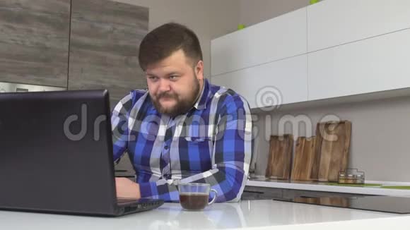 留胡子的男人在家里厨房里喝咖啡在笔记本电脑上工作在家工作自由职业者慢悠悠视频的预览图
