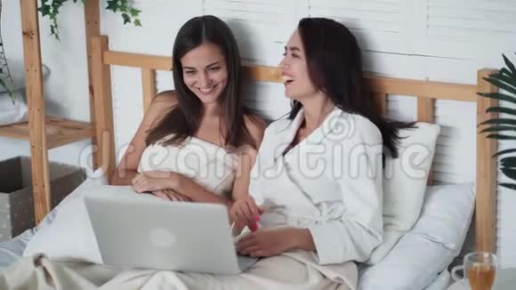 两个年轻的女朋友坐在床上在笔记本电脑上看滑稽电影笑着视频的预览图