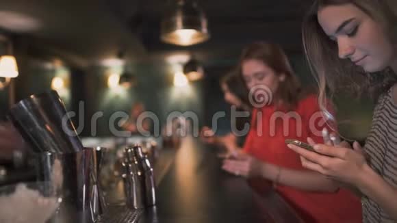 三个忙碌的年轻女孩坐在酒吧旁等待鸡尾酒在他们的手机上输入信息微笑着视频的预览图