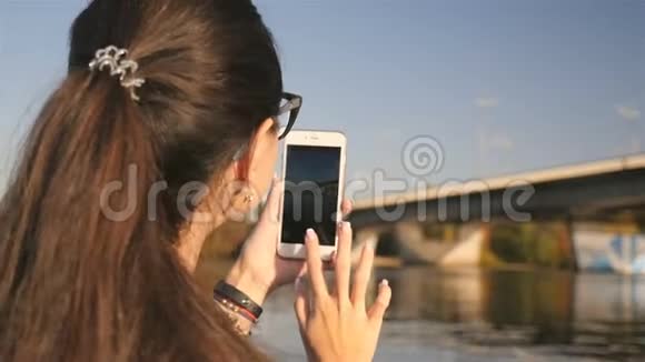一个年轻女孩在电话里拍照河上美丽的桥风景如画特写镜头晴天视频的预览图