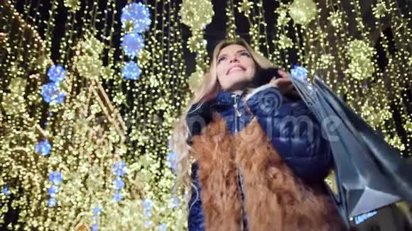 身穿毛皮大衣身穿圣诞购物袋身穿黄色灯光背景的迷人微笑女子视频的预览图
