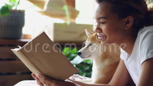 美籍美籍聪明的女性在空闲时间与她的带着大狗的家犬躺在床上看书的特写镜头视频的预览图