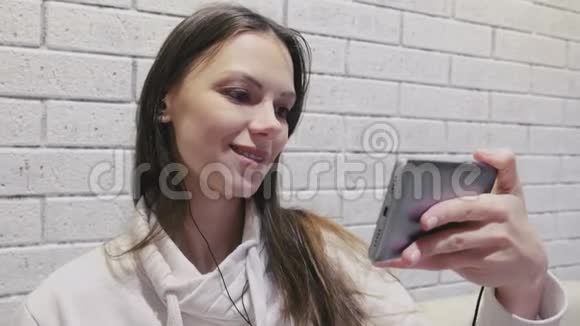 一位美丽的黑发女人在一堵砖白色的墙上看着一串戴着耳机的浪漫塞尔维亚剧视频的预览图