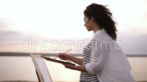 美丽的卷发女孩用颜料和画笔在湖边的草地上画一幅画采购文件视频的预览图