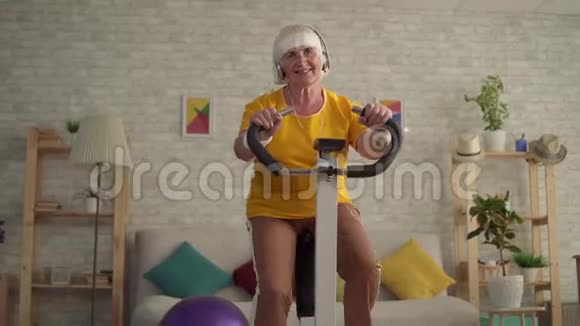 戴着运动自行车进行有氧训练的耳机的活跃老年妇女视频的预览图