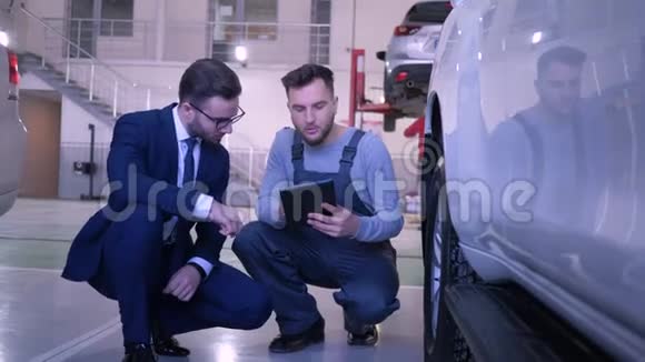 汽车维修厂汽车车轮附近的消费者与平板电脑的男技师沟通视频的预览图