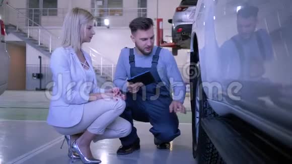 带平板电脑的机械师与加油站汽车轮附近的女性客户沟通视频的预览图