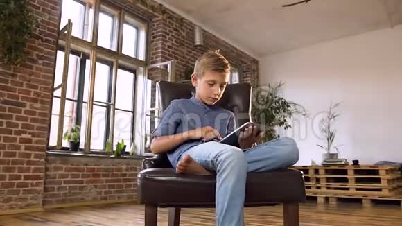 可爱的白种人男孩坐在扶手椅上在平板电脑上看有趣的电影一个严肃的少年男孩视频的预览图