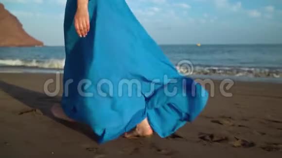 一个穿着漂亮蓝色裙子的女人的腿沿着黑色的火山海滩散步慢动作视频的预览图