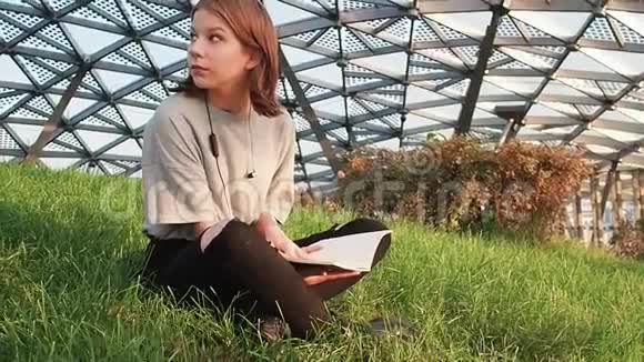 穿着灰色衬衫的少女坐在公园的草地上环顾四周在阳光明媚的阳光下看着一本笔记本视频的预览图