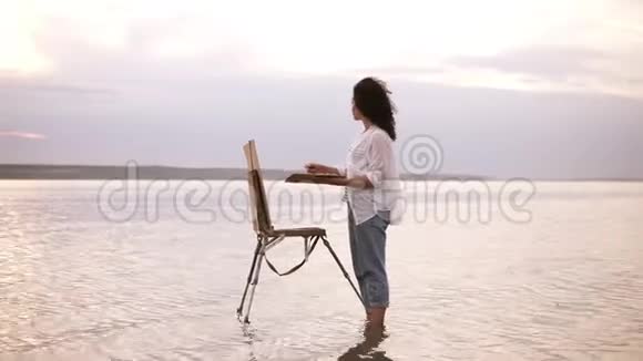 美丽的景色一个年轻的卷曲的黑发女子站在水中画架和她周围的景观视频的预览图