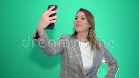 穿着夹克的快乐金发女人在绿色背景下的智能手机上自拍视频的预览图