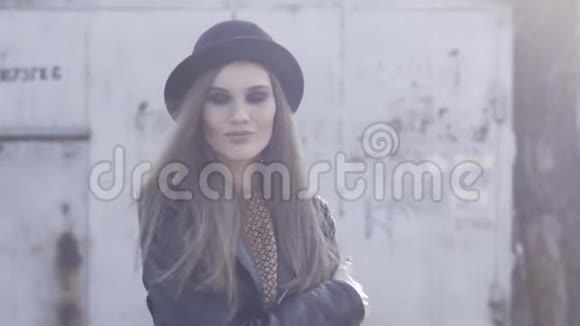 漂亮的时髦女孩带着黑色时髦的帽子和皮夹克在温暖的阳光下摆在老大楼里拥抱自己视频的预览图