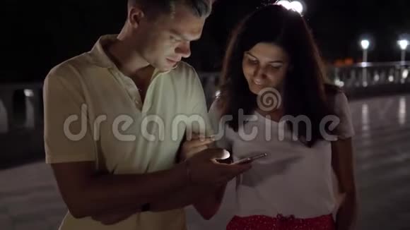 晚上快乐的一对情侣在街上一起看着智能手机视频的预览图