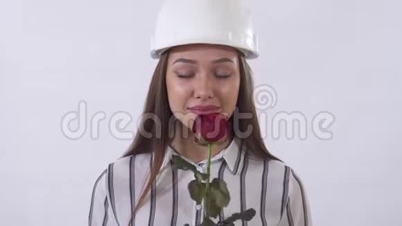 戴着建筑帽的年轻漂亮女人手里拿着一朵红玫瑰很高兴在画室里用白色拍摄视频的预览图