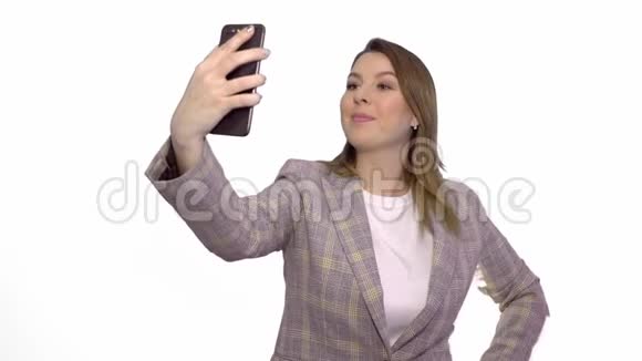 穿着夹克的快乐金发女人在白色背景的智能手机上自拍视频的预览图