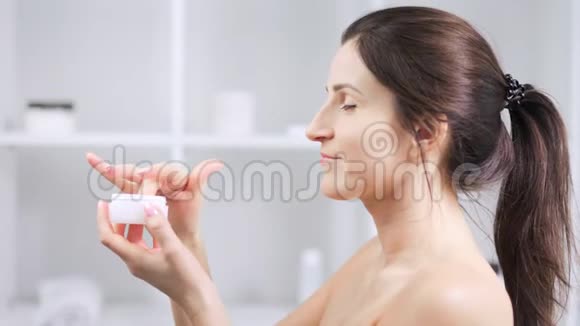 微笑的女人在白色浴室灯光背景下享受颈部保湿霜视频的预览图