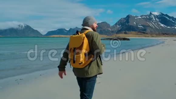 徒步旅行者穿着绿色夹克走在遥远的沙滩上背着一个带着大山的黄色背包破浪前行视频的预览图