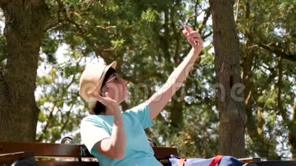 戴着帽子和太阳镜的快乐女孩坐在公园的长凳上打电话给自己拍照视频的预览图