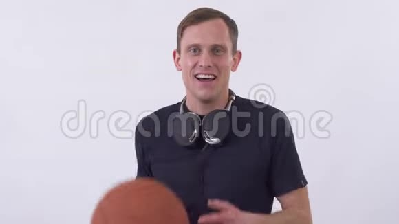 一个带着耳机的年轻人手里拿着篮球的画像在摄影棚拍摄白色背景视频的预览图
