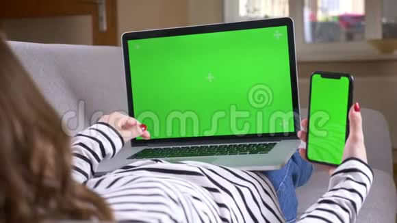 特写后景拍摄女性在笔记本电脑上打字和使用手机与绿色屏幕坐在沙发上视频的预览图