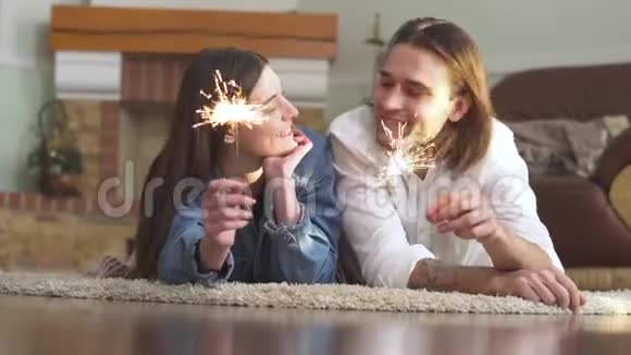 快乐的年轻美丽的男人和女孩手里拿着火花躺在家里的地毯上视频的预览图