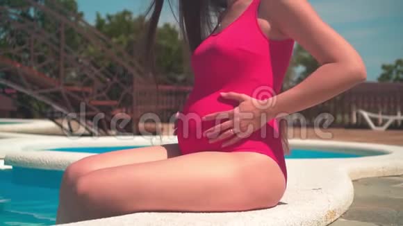 怀孕的女孩穿着红色的一件泳衣和眼镜坐在游泳池附近抚摸着她的肚子视频的预览图