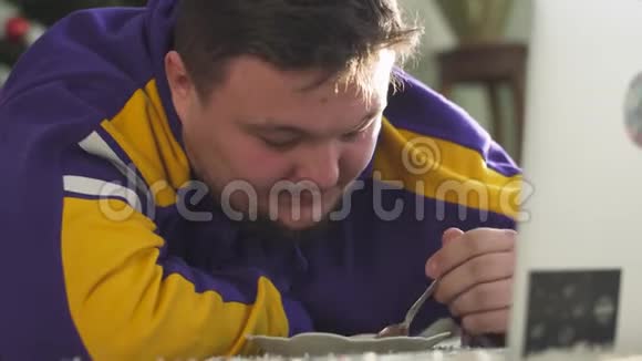 一个满脸胡须的年轻人坐在椅子上看着笔记本电脑用勺子吃果酱的肖像视频的预览图