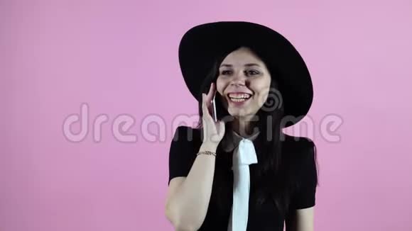 穿着黑色连衣裙和帽子的快乐女人在粉色背景的智能手机上交谈视频的预览图