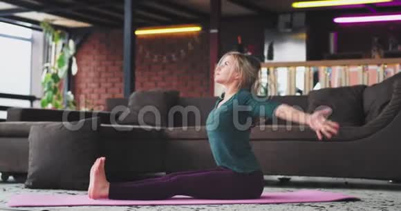 为一位穿着舒适运动服的金发女人在家做瑜伽冥想并在客厅练习瑜伽视频的预览图