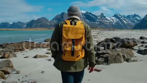 徒步旅行者穿着绿色夹克走在遥远的沙滩上背着一个带着大山的黄色背包破浪前行视频的预览图