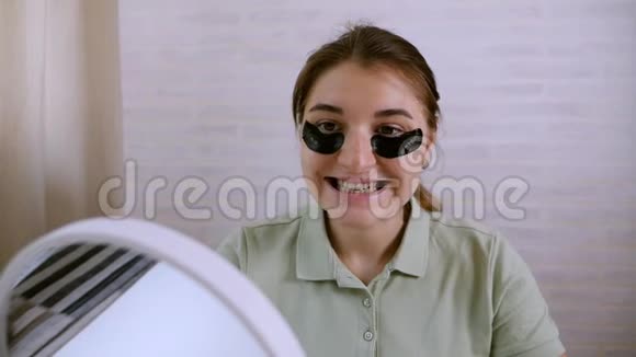 女孩用黑色补丁在她的眼睛下微笑坐在镜子对面健康的生活方式青春面部视频的预览图