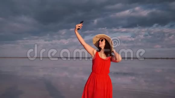 戴着帽子和太阳镜的年轻女人在天空和云彩的背景下自拍旅行者在电话里拍照视频的预览图