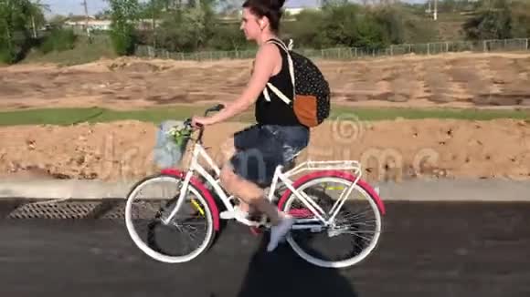 女孩骑自行车在沥青路上在她的背包后面在一个货篮里放着一束花视频的预览图
