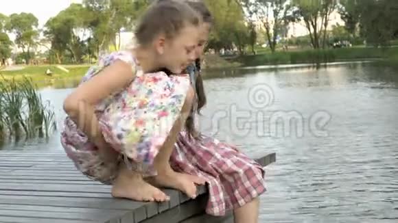 一个夏天的傍晚两个可爱的小女孩正坐在河岸上的一座木桥上肖像关门视频的预览图