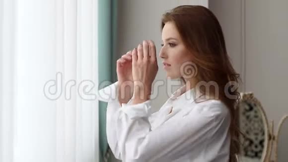 穿着白色衬衫的性感红发女孩站在卧室的窗户附近视频的预览图