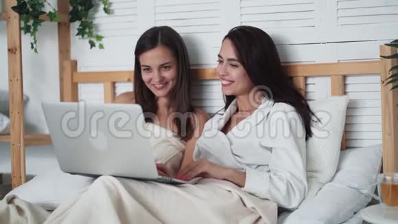 两个年轻的女朋友坐在床上在笔记本电脑上看滑稽电影笑着视频的预览图