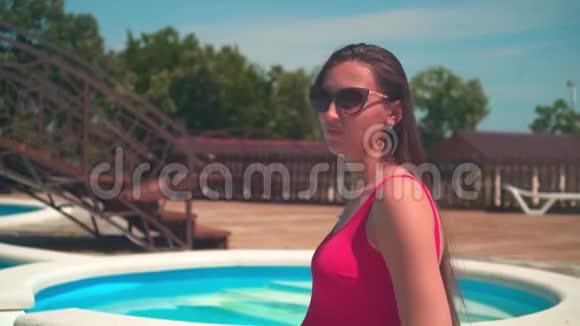 怀孕的女孩穿着红色的一件泳衣和眼镜坐在游泳池附近抚摸着她的肚子视频的预览图