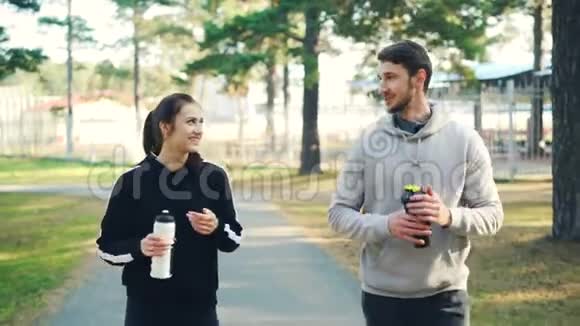 年轻夫妇穿着运动服手里拿着水瓶在森林里散步谈笑风生视频的预览图