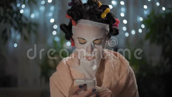 特写镜头穿着粉色长袍的女人在智能手机上写着一条信息用一个振兴面具和卷发器视频的预览图
