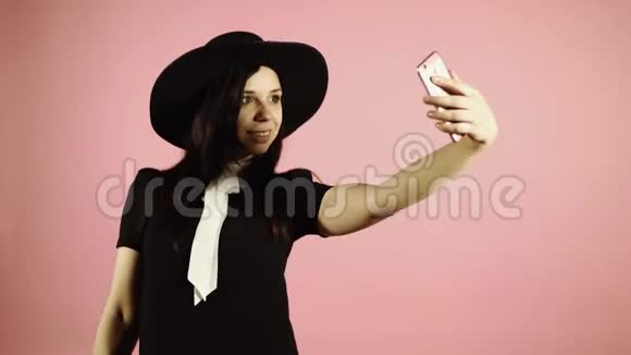 女士在智能手机上自拍穿着黑色连衣裙和帽子的女人带着粉红色背景的电话视频的预览图