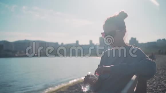 一个穿着运动衫和眼镜的黑发女孩坐在靠近大海的长凳上从塑料瓶里喝水视频的预览图
