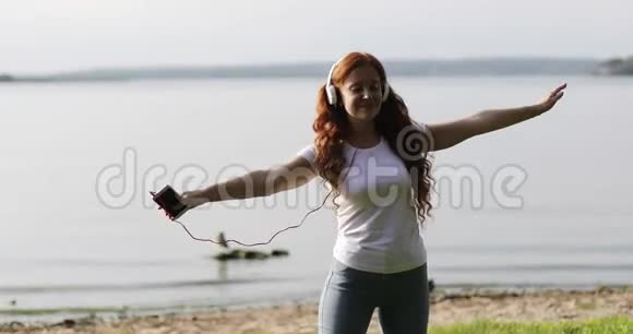 年轻快乐的女人红色卷发在湖岸的耳机里欣赏音乐视频的预览图